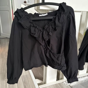 Svart blus med volang från NA-KD - Svart blus från NA-KD med bred volang längs halsringningen och omlott framtill. Långärmad modell med lätt puff vid axlarna och resår i midjan baktill. Tillverkad i mjuk bomull som känns skön mot huden. Tröjan är i storlek 40 men skulle säga att den är mer som en 36/38. 