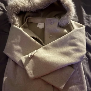 Beige kappa med fuskpäls från Gina Tricot - Jättefin och skön beige kappa från Gina Tricot köptes för en månad sen. med fluffig fuskpäls runt huvan. Klassisk rak modell med knappar framtill och två fickor. Perfekt för dig som vill ha en stilren look med lite extra cozy känsla. Passar dig som gillar neutrala färger och mjuka material. I storlek 158-164strl 💗
