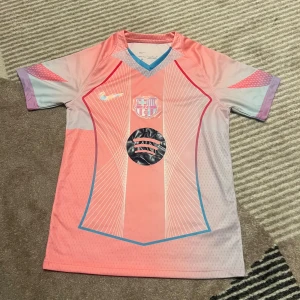 Rosa FC Barcelona fotbollströja Nike - Säljer en unik FC Barcelona fotbollströja från Nike i rosa och ljuslila med korta ärmar. Tröjan har klubbmärke och sponsortryck på bröstet samt grafiska linjer i vitt och blått. Materialet är lätt och andas, perfekt för träning eller match.