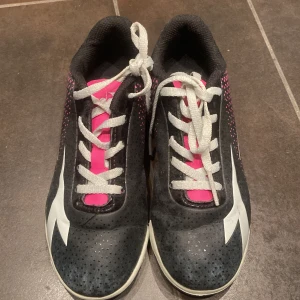 Diadora svarta och rosa fotbollsskor - Säljer ett par Diadora fotbollsskor i svart och rosa med vita detaljer. Skorna har snörning, perforerat syntetmaterial och mönstrad sula för bra grepp på planen. Snygg design med grafiskt mönster på sidorna och tydlig Diadora-logga. Perfekta för dig som vill sticka ut på fotbollsplanen.