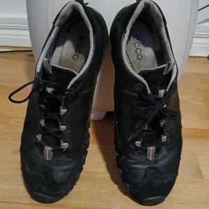 Svarta sneakers från ECCO i skinn - Svarta sneakers från ECCO med coolt mönstrad sula och snörning. Skorna är gjorda i skinn och har detaljer i grått och lila på plösen och snörningen. Perfekta för dig som gillar en sportig och stilren look. Passar lika bra i hösten.