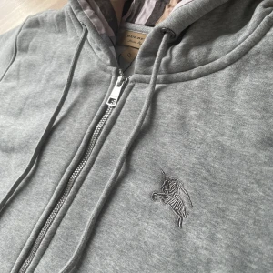 Burberry Zipup - Snygg grå hoodie från Burberry med klassiskt rutigt foder i huvan. Tröjan har dragkedja framtill, snörning i huvan och broderad logga på bröstet. Tillverkad i mjuk bomullsmix, perfekt för chill dagar. Stora fickor framtill och ribbade muddar.