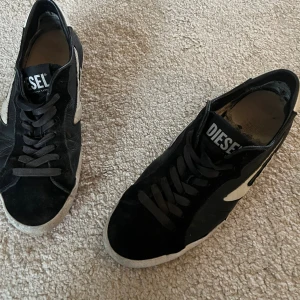 Svarta sneakers från Diesel - Svarta sneakers från Diesel med vita detaljer på sidorna och vit sula. Skorna har snörning och en röd undersida på sulan som ger en cool kontrast. Ovandelen är i skinn och mocka, med Diesel-logga på plösen. Perfekta för en streetwear-look. OBS! Inte bästa skicket, synns dem två sista bilderna, sulan är dålig och kanske tillfälle att byta och det är en lite spricka på yttersida, därav priset. Priset är ej hugget i sten! 