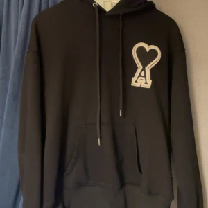 Svart hoodie från Ami med hjärtdetalj - Svart hoodie från Ami Alexandre Mattiussi med stor vit hjärtdetalj och bokstaven A på bröstet. Tröjan har huva med snörning, känguruficka och ribbade muddar. Tillverkad i mjuk bomull, perfekt för chill dagar. Snygg och stilren design som sticker ut. Pris kan justeras vid snabb affär 