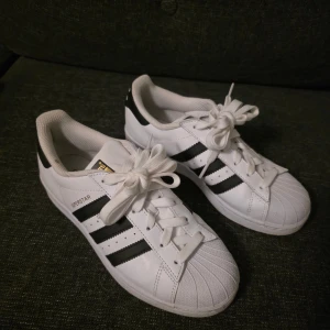 Adidas Superstar sneakers vit/svart 37⅓ - Klassiska Adidas Superstar sneakers i vitt skinn med svarta ränder och detaljer. Ikonisk snäcktå, platt sula och snörning framtill. Guldiga detaljer på plösen och Adidas-logga bak. Perfekta för dig som gillar streetstyle och tidlös design.