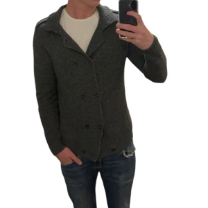 Mörkgrå stickad kofta med krage - Snygg mörkgrå stickad kofta med bred krage och dubbelknäppning framtill. Koftan har långärmade ärmar och en grovstickad struktur som ger en cool och avslappnad vibe. Perfekt att slänga över en t-shirt för en enkel men stilren look.