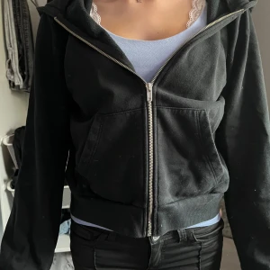 Svart hoodie med dragkedja och huva - Svart hoodie med huva och silverfärgad dragkedja framtill. Tröjan har långa ärmar, ribbade muddar och en stor ficka på magen. Perfekt för chill dagar och enkel att matcha med jeans eller leggings.