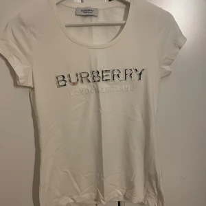 Vit Burberry t-shirt - Vit t-shirt från Burberry med broderad logga i silver och svart på bröstet. Klassisk rund halsringning och korta ärmar. Tillverkad i mjuk material för en skön känsla. Snygg och stilren design med exklusiv känsla.