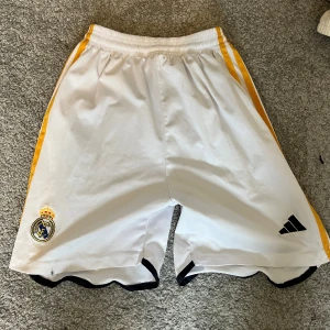 Real Madrid vita fotbollsshorts Adidas - Snygga vita fotbollsshorts från Adidas med Real Madrid-logga på ena benet och tre gula ränder längs sidorna. Svarta detaljer vid benslut och elastisk midja för skön passform. Perfekta för dig som älskar fotboll och vill ha en sportig look.