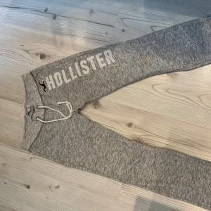 Grå lågmidjade mjukisbyxor Hollister XS - Snygga lågmidjade grå mjukisbyxor från Hollister i storlek XS med vit snörning i midjan och stort broderat Hollister-tryck längs benet. Mjukt material och avslappnad passform, perfekta för chill eller träning. Klassisk logga i vinrött vid höften.