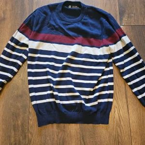 Randig blå tröja från Albion XL - Snygg långärmad tröja från Albion i storlek XL. Tröjan har marinblå och vita ränder med en vinröd detalj över bröstet. Rund hals och mjukt material som känns skönt mot huden. Perfekt för dig som gillar klassisk stil med en twist.