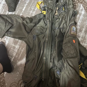 Grön parkajacka från Parajumpers - Säljer en grön Parajumpers parkajacka med huva, coola patchar på ärmen och detaljer i gult. Jackan har flera fickor, dragkedja och knappar framtill. Perfekt för dig som vill ha en snygg och funktionell jacka med street-vibe.