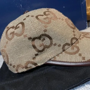Gucci beige keps med GG-mönster - Snygg beige keps från Gucci med det klassiska GG-mönstret i brunt över hela kepsen. Kepsen har böjd skärm med brunt kantband och är tillverkad i ett slitstarkt tygmaterial. Perfekt för dig som vill ha en lyxig och trendig accessoar till din outfit.