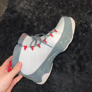Nike Jordan 9 Retro vit/grå/röd - Nike Jordan 9 Retro sneakers i vitt skinn med grå och röda detaljer. Skon har hög siluett, snörning med röda öglor och grå sula i blank finish. Ikonisk design med mönstrade paneler och bekväm passform. Perfekt för dig som gillar streetwear och basketstil.