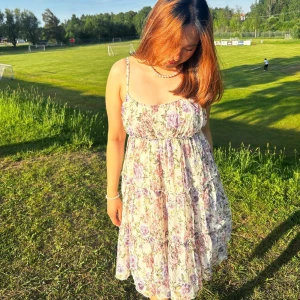 Blommig sommarklänning med volanger - Supersöt blommig klänning i ljusa toner med lila och rosa blommor. Klänningen har smala axelband, volanger och en luftig passform som passar perfekt för varma dagar. Materialet känns lätt och flowy, perfekt för sommarens alla äventyr.