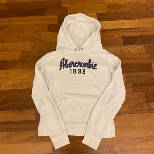 Vit hoodie från Abercrombie & Fitch - Vit hoodie från Abercrombie & Fitch med broderad logga och årtal på bröstet. Klassisk känguruficka framtill, huva med snörhål och långa ärmar. Mjuk insida och avslappnad passform, perfekt för chill dagar. Passar till en liten s