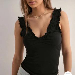 Svart volangärmad linnetopp - Snygg svart linnetopp med djup v-ringning och breda axelband med volangdetaljer. Toppen är figurnära och har en feminin siluett, perfekt att styla med jeans eller kjol. Materialet känns mjukt och stretchigt mot huden.