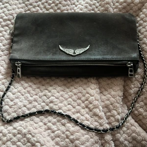 Svart axelväska Zadig & Voltaire skinn - Cool svart axelväska i skinn från Zadig & Voltaire med silverfärgad kedjerem och ikonisk vinge framtill. Väskan har en dragkedja och detaljer i silvermetall. Perfekt storlek för det viktigaste och ger en edgy vibe till din outfit.