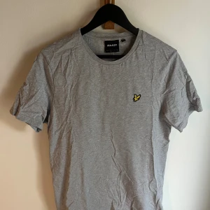 Grå t-shirt från Lyle & Scott - Snygg grå t-shirt från Lyle & Scott med klassisk rund hals och korta ärmar. På bröstet sitter den ikoniska gula fågeln som broderad detalj. T-shirten är gjord i mjuk bomull och har en clean, stilren look som funkar till allt.