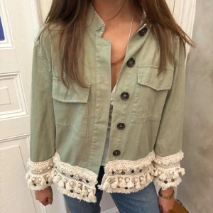 Grön jeansjacka med detaljer - Snygg ljusgrön overshirt med stora svarta knappar och två bröstfickor. Jackan har unika virkade detaljer och tofsar i beige längs ärmslut och nederkant. Perfekt för dig som vill sticka ut med en bohemisk touch. Materialet känns som bomull och passformen är loose.
