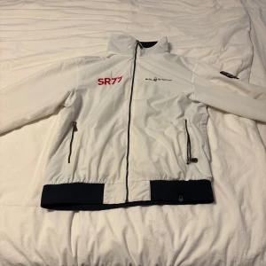 Vit vindjacka Sail Racing SR77 - Vit vindjacka från Sail Racing med SR77-tryck och broderad Extreme Sailing-logga. Jackan har hel dragkedja, höga muddar i mörkblått, två fickor med dragkedja och meshfoder. Snygga detaljer med flaggor och text på ryggen. Perfekt för dig som gillar sportig stil.