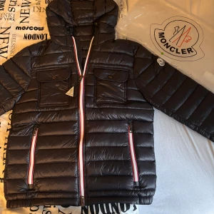Svart Moncler dunjacka med huva - Svart dunjacka från Moncler, huva och två stora bröstfickor med lock. Jackan har röda och vita detaljer på dragkedjorna och klassisk Moncler-logga på ärmen. Perfekt för kalla dagar och riktigt snygg streetstil. Det är storlek 1 motsvarar S och passar någon som är ca 177cm lång och väger 65kg. Finns ingen i Sverige som säljer detta skicket på jackan för ett så billigt pris. INGA HÅL ELLER SKADOR.