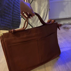 Brun portfölj i skinn med dragkedja - Snygg portfölj i brunt skinn med två stora fickor framtill, guldfärgade detaljer och dragkedja. Väskan har både handtag och axelrem, vilket gör den enkel att bära. Perfekt för laptop, böcker eller dokument. Stilren och klassisk design.