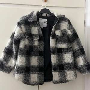 Rutig teddyjacka från Skill, svart/vit - Mysig teddyjacka från Skill i svart och vitt rutigt mönster. Jackan har stora knappar framtill, två bröstfickor med lock och klassisk krage. Perfekt för kalla dagar när du vill ha en cool och avslappnad stil.