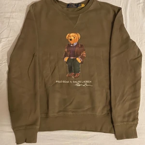 Olivgrön Polo Bear tröja Ralph Lauren - Olivgrön crewneck från Polo Ralph Lauren med ikoniska Polo Bear-trycket framtill. Tröjan har rund hals, långa ärmar och ribbade muddar. Perfekt för dig som gillar klassisk streetstyle med en twist. Materialet är mjuk bomull som känns skönt mot huden.