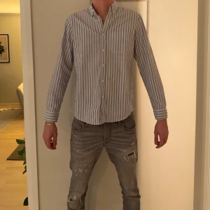 Randig skjorta från Pull&Bear M - Snygg randig skjorta från Pull&Bear i storlek M. Skjortan har vita och gråa ränder, klassisk krage, bröstficka och knäppning framtill. Regular fit och långärmad, perfekt för en clean och stilren look.