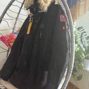 Svart Parajumpers vinterjacka med päls - Grym svart vinterjacka från Parajumpers med ikonisk patch på ärmen, gul detalj vid kragen och fet päls runt huvan. Jackan har flera fickor, dragkedja och knappar framtill. Perfekt för kalla dagar och riktigt snygg streetstil.