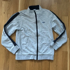 Grå ziptröja från BOSS med blå detaljer - Snygg grå ziptröja från BOSS med svarta och blå ränder längs ärmarna. Tröjan har hög krage, två fickor fram och svart dragkedja. Perfekt för chill eller sportig stil, med diskret BOSS-logga på bröstet. Mjuk bomullskänsla och normal passform. Kan gå ner i pris