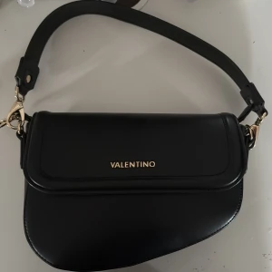 Svart axelväska från Mario Valentino - Snygg svart axelväska från Mario Valentino med stilren design och guldfärgade detaljer. Väskan har ett avtagbart axelband samt extra lång rem för olika stilar. Perfekt storlek för det viktigaste och har dragkedja på insidan. Väskan är även i nyskick. 