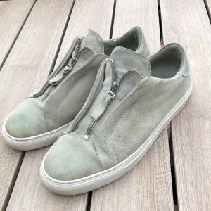 Ljusgrå  med dragkedja - Snygga arigato skor i ljusgrå i mocka med vita sulor och dragkedja framtill istället för klassisk snörning. Skorna har rund tå och detaljer i ljusgrått skinn vid hälen. Perfekta för dig som gillar stilrena och minimalistiska skor med en twist. Inga defekter förutom en liten fläck. (Se bild 3) skick 8/10. hör av er vid frågor!🙏🏻