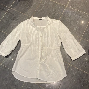 Vit skjorta med knappar från Esmara - Vit skjorta från Esmara i bomull med snygga veckade detaljer framtill och knappar längs hela framsidan. Skjortan har trekvartsärm och en lätt loose passform med dragsko i midjan för en chill look. Perfekt för dig som gillar enkel och stilren design.