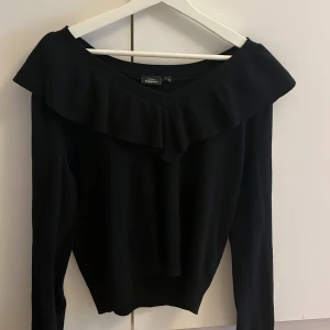 Svart volangtröja från KappAhl - Svart långärmad tröja från KappAhl med bred volang längs halsringningen. Tröjan har en snygg v-ringad design och är gjord i mjukt stickat material. Perfekt för dig som gillar stilrena och trendiga detaljer.