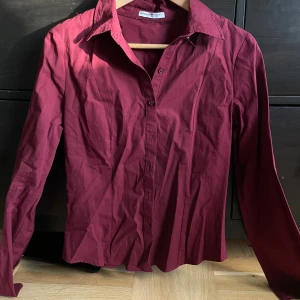 Vinröd figursydd skjorta med knappar - Vinröd skjorta från bershka i klassisk modell med lång ärm och krage. Skjortan har en figurnära passform och stängs med matchande knappar framtill. Materialet är mjukt och känns som bomull, perfekt för dig som gillar stilrena plagg med enkel design. Aldrig använd. Kom med en knapp som var sönder