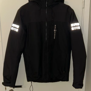 Svart vinterjacka från Sail Racing - Svart winterjacka från Sail Racing med reflekterande detaljer på ärmarna och logga på bröstet. Jackan har huva med snörning, dragkedja framtill och justerbara ärmslut. Perfekt för kalla dagar och snygg streetstil. Köpte den för ungefär 3000 kr. Helt ny bestälde den men fel storlek, kunde it skicka tillbaka den så försöker sälja den här nu .