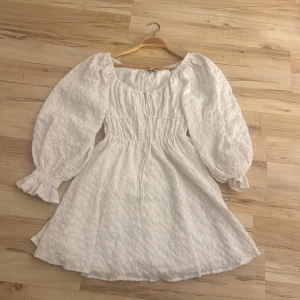 Vit kort klänning med puffärmar - Supersöt vit kort klänning med puffiga långärmar och smock i midjan. Klänningen har en lätt textur i tyget och en rundad halsringning. Perfekt för dig som gillar romantisk och trendig stil med volym och detaljer.