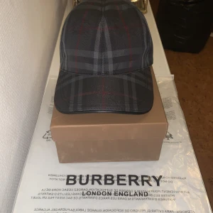 Rutig keps från Burberry i mörka toner - Snygg keps från Burberry i klassiskt rutigt mönster med mörkblå, grå och röda detaljer. Kepsen har böjd skärm och justerbar passform. Perfekt accessoar för att lyfta din streetstyle med exklusiv känsla. Kommer med originalbox och tag.
