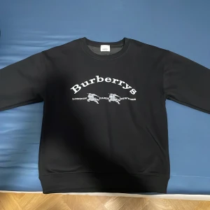 Svart sweatshirt från Burberry - Svart sweatshirt från Burberry med klassisk rund hals och tryck i vitt framtill med texten 'Burberrys' och motiv. Tröjan har långa ärmar och ribbade muddar vid ärmslut och nederkant. Perfekt för en stilren och trendig look.. Kan gå ner i pris