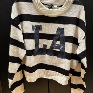 Randig stickad tröja från H&M Divided - Snygg stickad tröja från H&M Divided i svart och vitt med breda ränder och tryckt LA-text framtill. Tröjan har rund halsringning, långa ärmar och en avslappnad passform. Perfekt för dig som gillar streetstyle och vill ha en cool look.