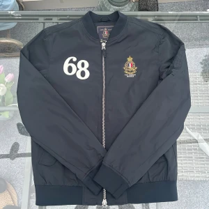 Morris Gents bomberjacka navy stl S - Snygg och stilren Morris Gents bomberjacka i mörkblått (navy). Klassisk modell med dragkedja, ribbade muddar och snyggt broderat Morris-emblem på bröstet samt siffra “68” på höger sida. Insidan har ikoniskt Union Jack-foder i rött, vitt och blått – en riktig statement-detalj. Storlek Small, passar true to size. Perfekt till både vardag och fest. I fint skick, endast sparsamt använd.  💙 Detaljer: 	•	Färg: Navy / mörkblå 	•	Storlek: S 	•	Märke: Morris Gents 	•	Foder med brittisk flagga 	•	Material
