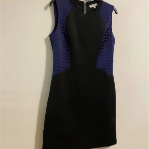 Svart och Lila  cocktail klänning från Sandro - Supersnygg svart kortklänning från Sandro med lila quiltade paneler på sidorna. Klänningen är ärmlös och har en rund halsringning samt dragkedja i ryggen. Materialet är mestadels polyester med detaljer i silke för en lyxig känsla. Perfekt för dig som gillar stilrena och moderna plagg.
