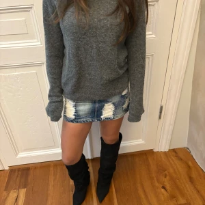 Blå jeanskjol med slitningar - Säljer en kort jeanskjol i blå denim med tydliga slitningar och fransiga detaljer både fram och bak. Kjolen har klassiska fickor, bälteshällor och en knapp framtill. Perfekt för dig som gillar en edgy och avslappnad stil.