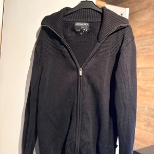 Svart stickad kofta Jack & Jones - Svart stickad kofta från Jack & Jones med grov ribbad krage och dragkedja framtill. Snygg struktur på axlar och nederkant, långärmad och perfekt för kyliga dagar. Enkel att matcha med jeans eller chinos.