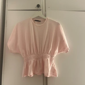 Ljusrosa blus med volang - Säljer en ljusrosa blus från lager 157 med korta ärmar och rund hals. Toppen har en snygg volang nedtill och smockad midja som ger en feminin siluett. Perfekt för dig som gillar söta och trendiga detaljer.