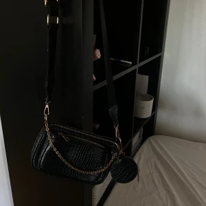 Svart axelväska med kedja - Snygg svart axelväska med croco-mönster och guldfärgad kedja. Väskan har flera dragkedjor och en rund liten plånbok som hänger på sidan. Justerbar axelrem i textil och detaljer i skinnimitation ger en cool vibe.