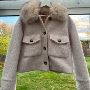 Pälskappa - Superfin beige kappa med stor fluffig päls på kragen. Jackan har två stora fickor framtill samt knappar. Perfekt för höst och vinter när du vill vara både varm och snygg. Det är två små fläckar på jackan som är inringade på bilderna! Den svarta är på baksidan av armen och den andra är typ vid knapparna så det är inget som syns jätte tydligt☺️