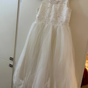 Vit långklänning för barn med spets och pärlor - Drömmig vit långklänning med blommig spets och pärldetaljer på överdelen. Kjolen är i flera lager tunn tyll med spetskant nertill och ett fint band i midjan som knyts bak. Öppen rygg med rund utskärning ger en extra elegant touch.